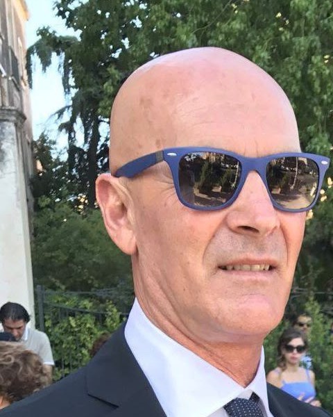 Uomo calvo con occhiali da sole blu a specchio, giacca scura, camicia bianca e cravatta a pois tra alberi e persone