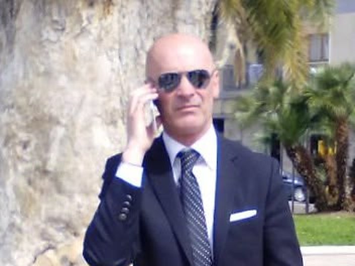 Uomo calvo con occhiali da sole, abito scuro e cravatta al telefono accanto a un grosso tronco, con palme sullo sfondo